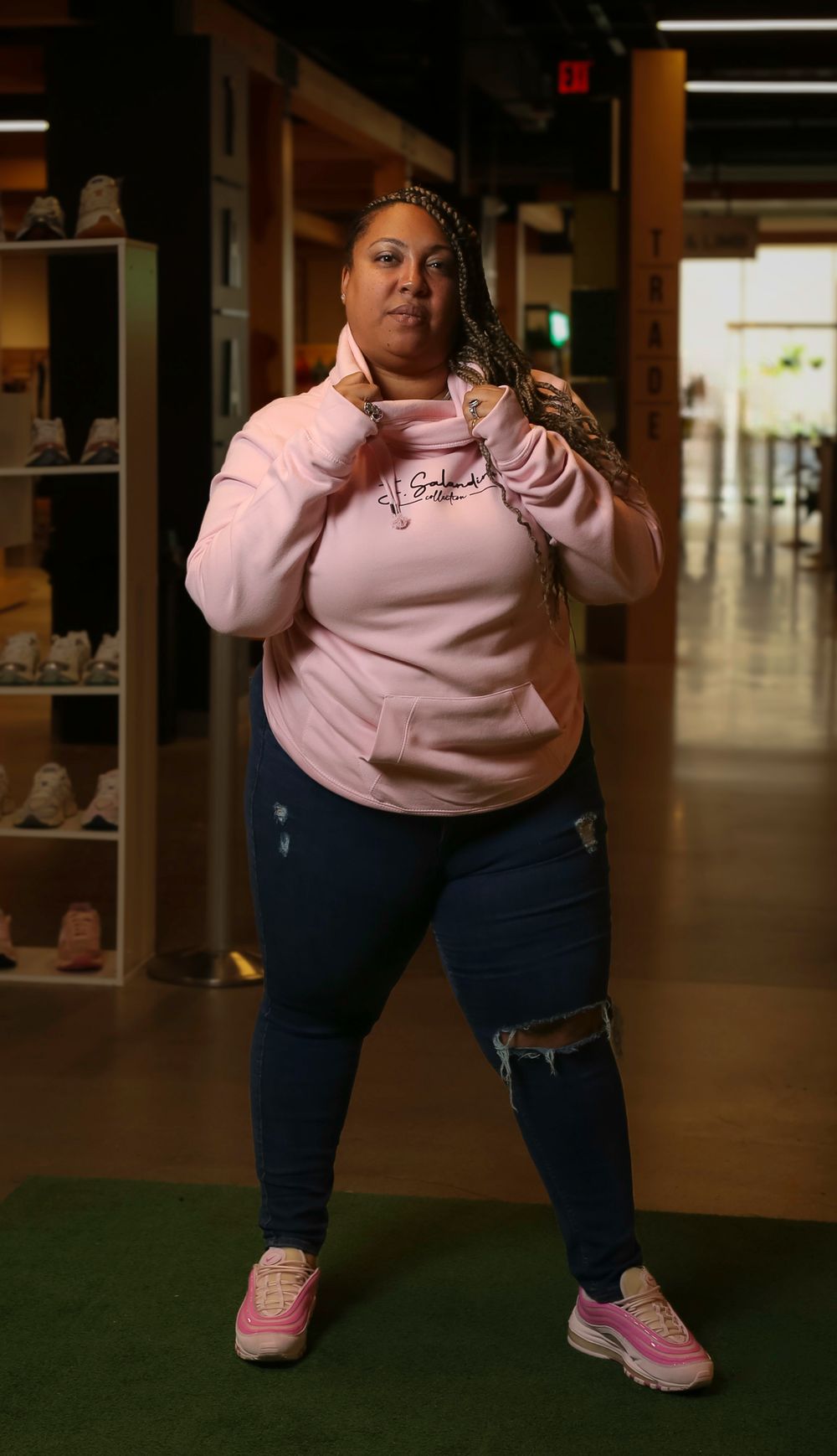 A person wearing a pink hoodie and ripped jeans poses confidently in a retail store with shoes displayed in the background.
