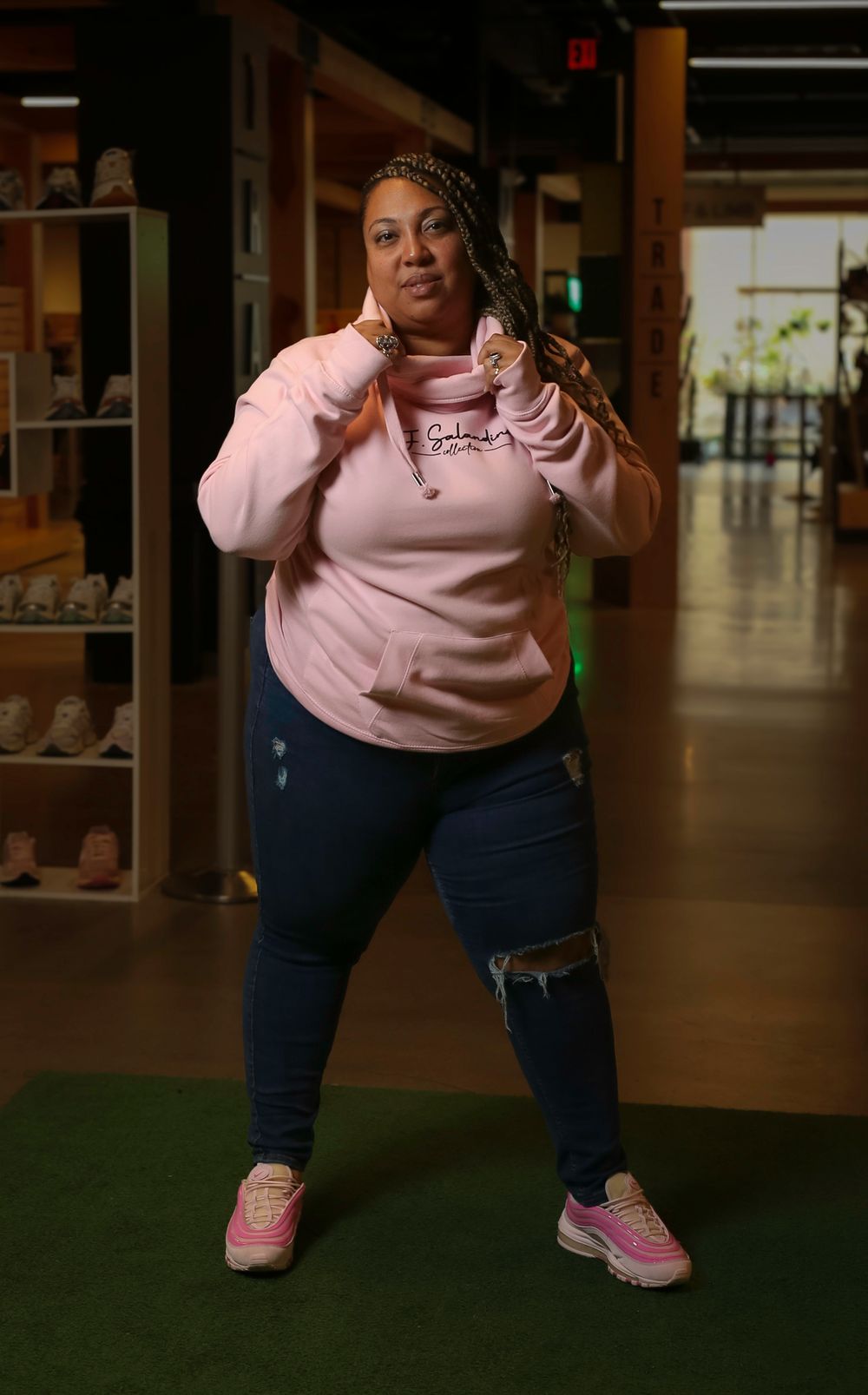 Person in pink sweatshirt and ripped jeans poses confidently in front of shoe display shelves.