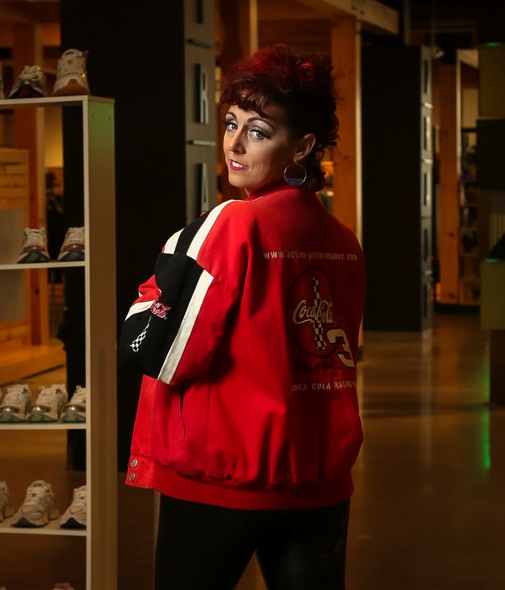 Person wearing a bright red varsity-style jacket poses in retail shop interior with shoe displays visible in background.