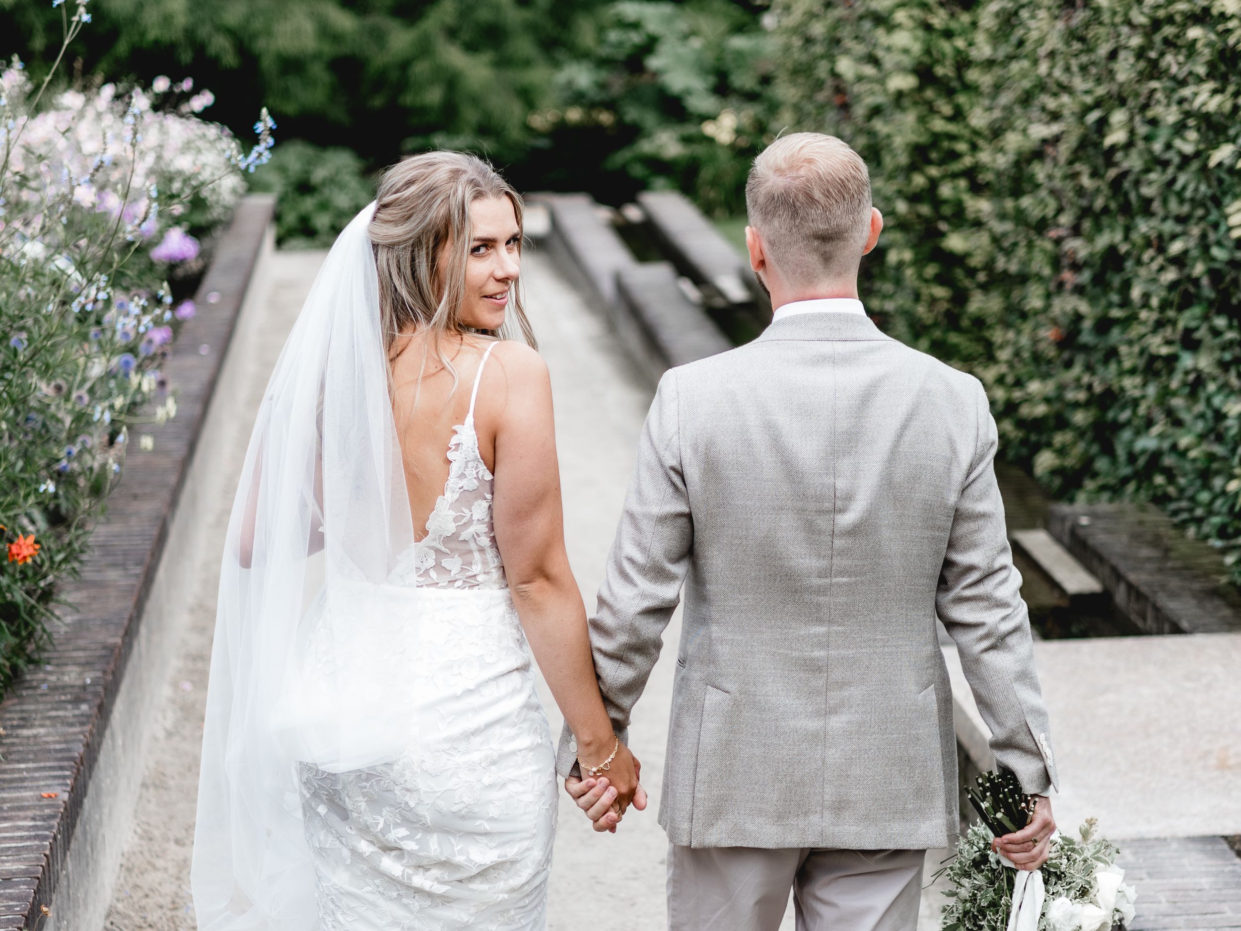 Couple walking through a lush garden pathway in wedding attire.