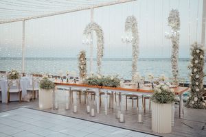 Beachfront wedding ceremony setup with white chairs, floral arrangements and draped fabric overlooking the ocean.