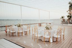 Elegant white beach wedding reception setup with round tables and chairs on a wooden deck overlooking the ocean.