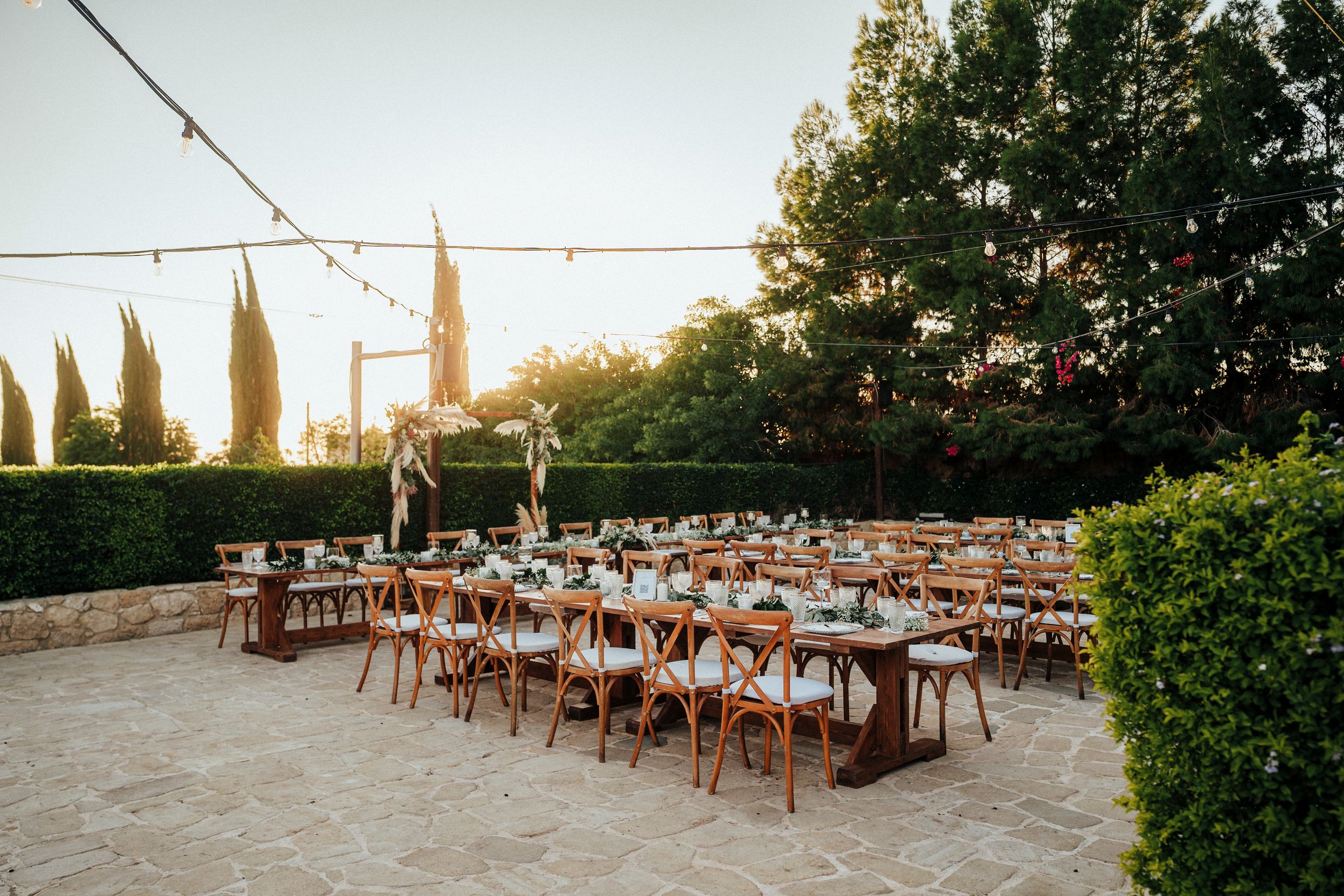 Rustic wooden chairs and tables set up for an outdoor wedding reception at sunset with string lights overhead.