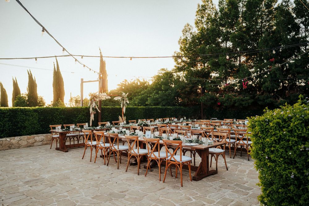 Rustic wooden chairs and tables set up for an outdoor wedding reception at sunset with string lights overhead.