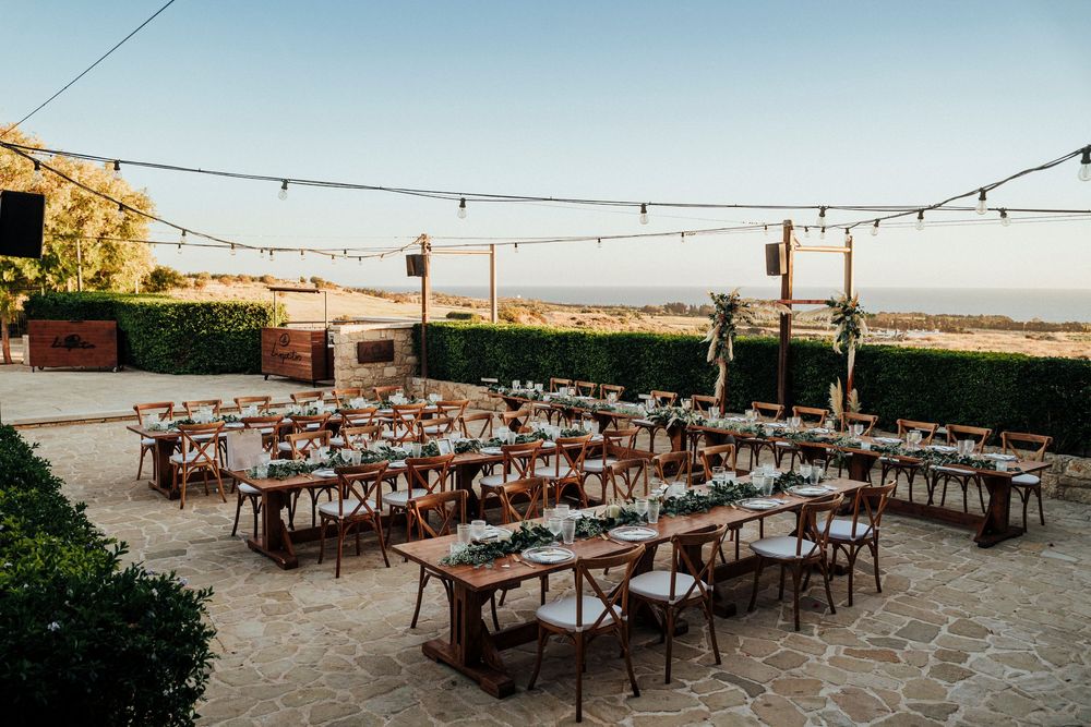 String lights illuminate an outdoor reception setup with rustic wooden tables and chairs arranged on a stone patio.