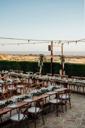 Elegant outdoor wedding reception setup with wooden tables and chairs on a stone patio overlooking desert mountains at sunset.