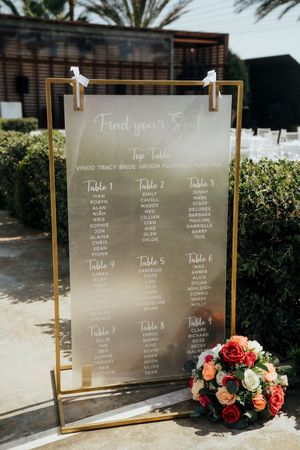 Modern wedding seating chart displayed on glass panels with gold frames and decorated with coral and peach floral arrangements.