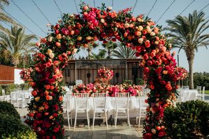 Panoramic view of a wedding ceremony setup with red floral arches and white chairs under palm trees.
