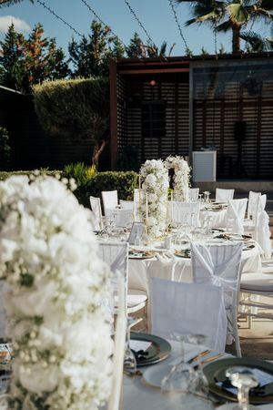 Elegant outdoor wedding reception tables decorated with white floral centerpieces and place settings under a pergola.