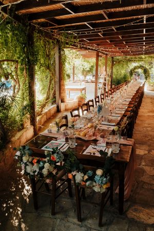 Long rustic wooden tables set up under a covered patio with string lights and floral centerpieces for an outdoor event.