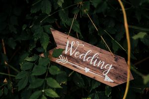 A rustic wooden wedding sign with white lettering and arrow design hangs among green foliage.