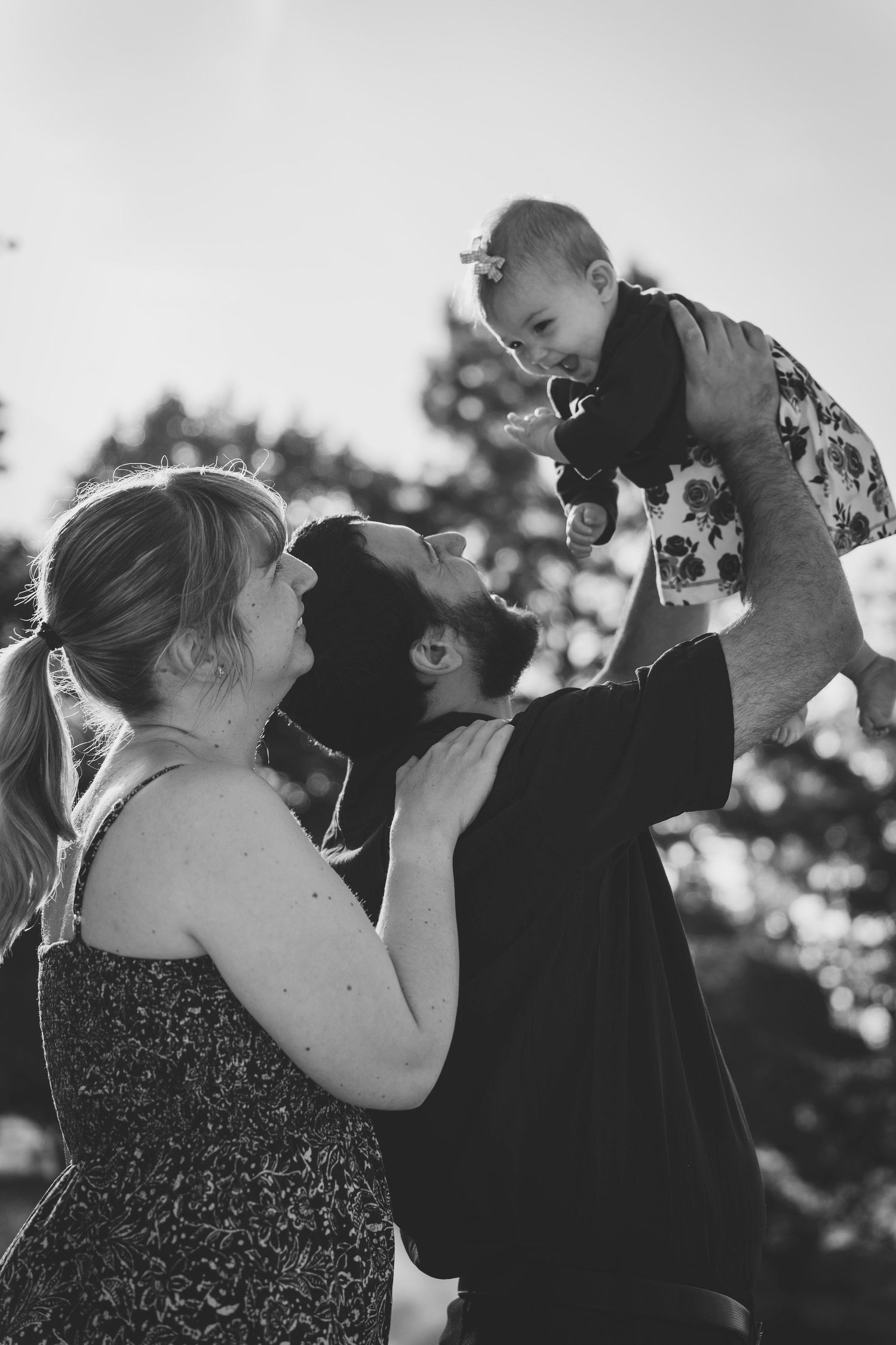 A joyful family moment captured in black and white as parents lift their small child high into the air outdoors.