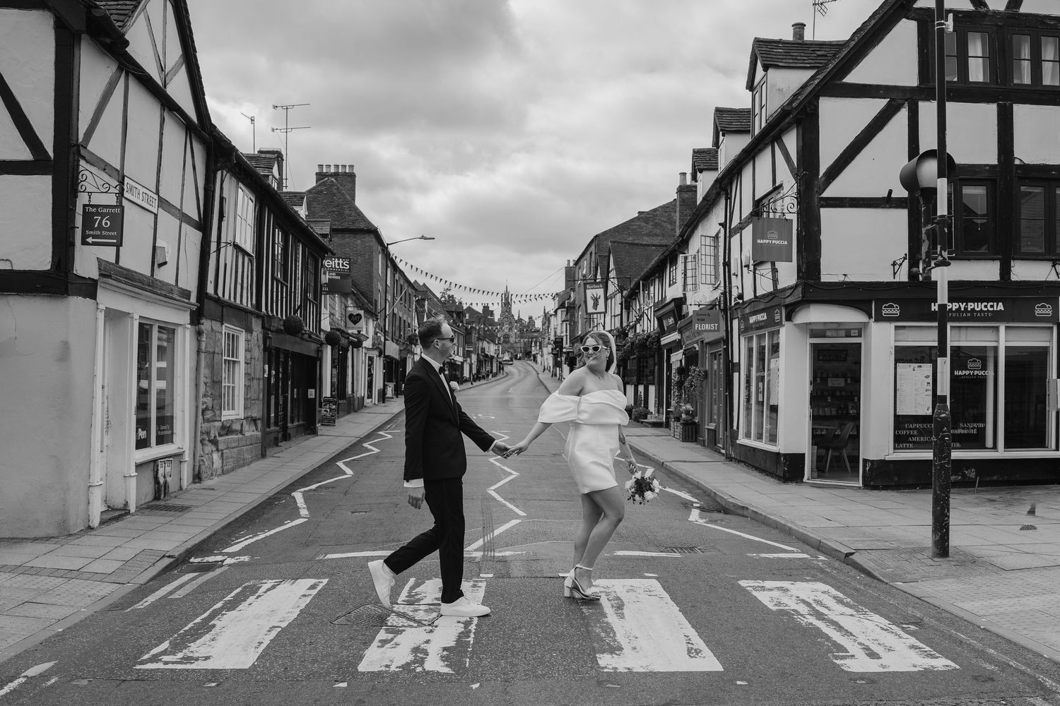 Black and white photo of a couple holding hands crossing a zebra crossing on a charming UK high street. warwickshire wedding photographer