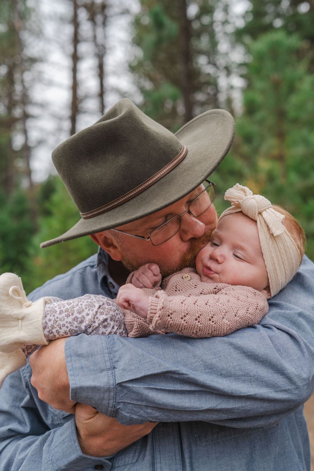A tender moment captured in nature as someone wearing a brown hat cradles a small baby in a pink sweater among pine trees.