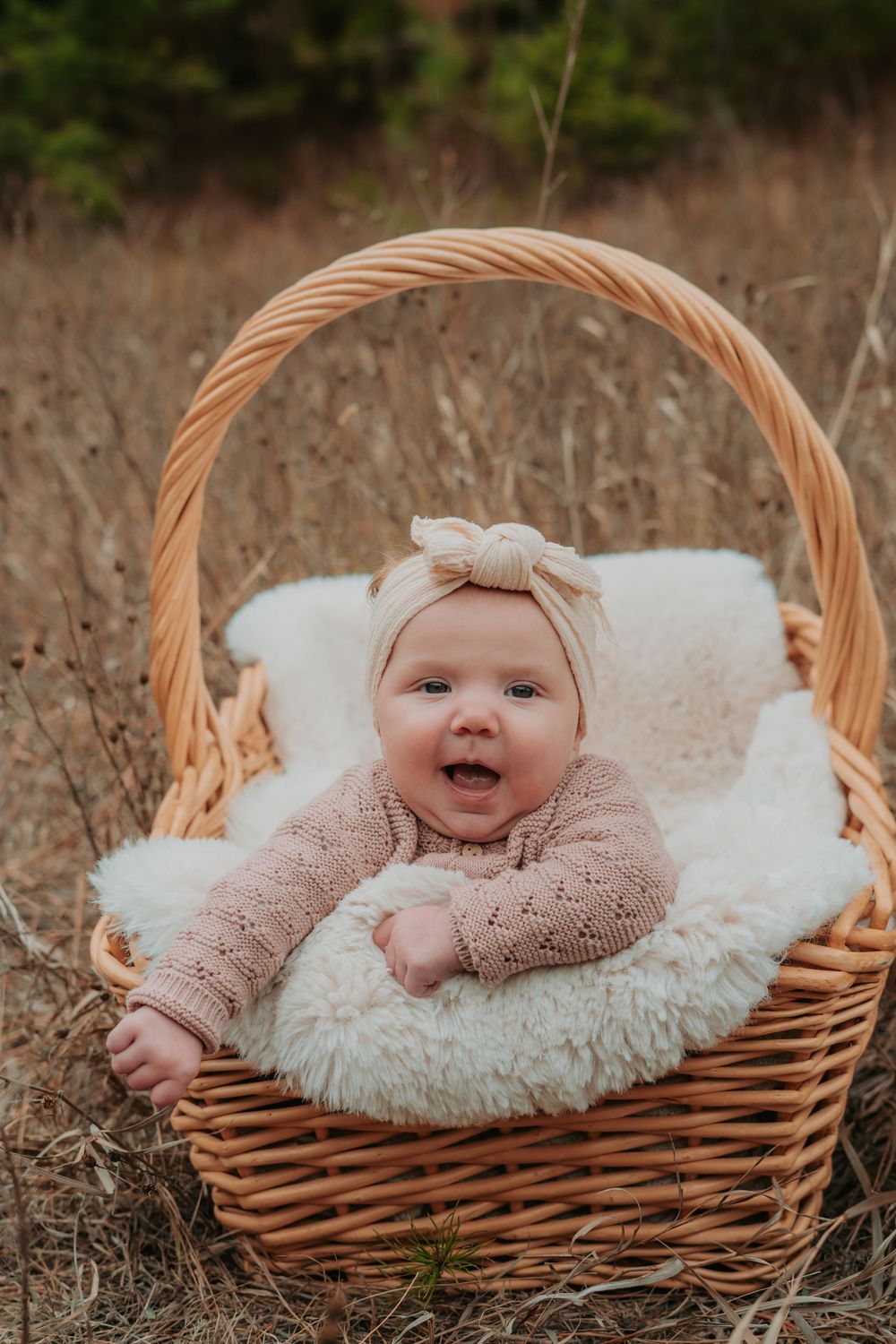 A baby lies in a woven basket with white fluffy sheepskin bedding during an outdoor photo session.