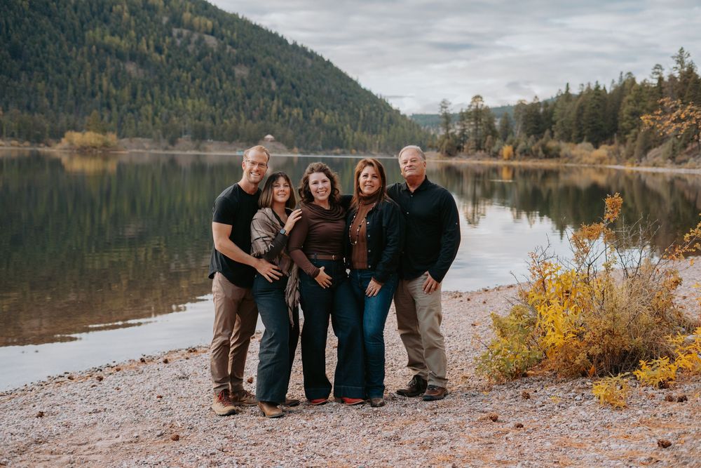 A group sits together on a rocky beach shore with mountains and a serene lake in the background during autumn.