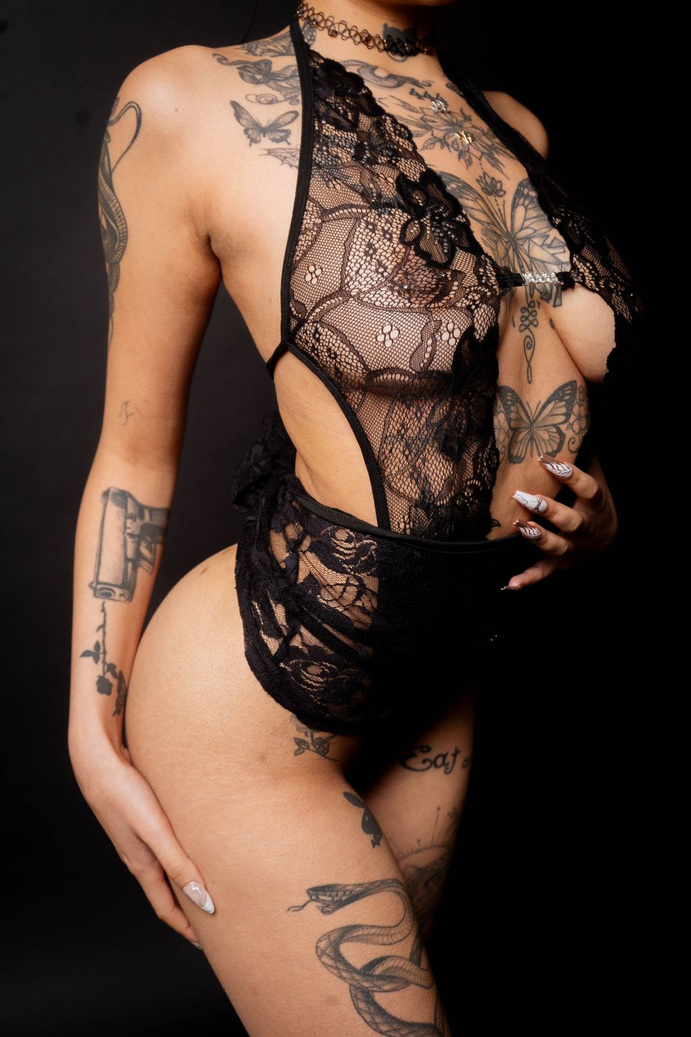 Heavily tattooed woman posing in black lace lingerie bodysuit against dark studio background.