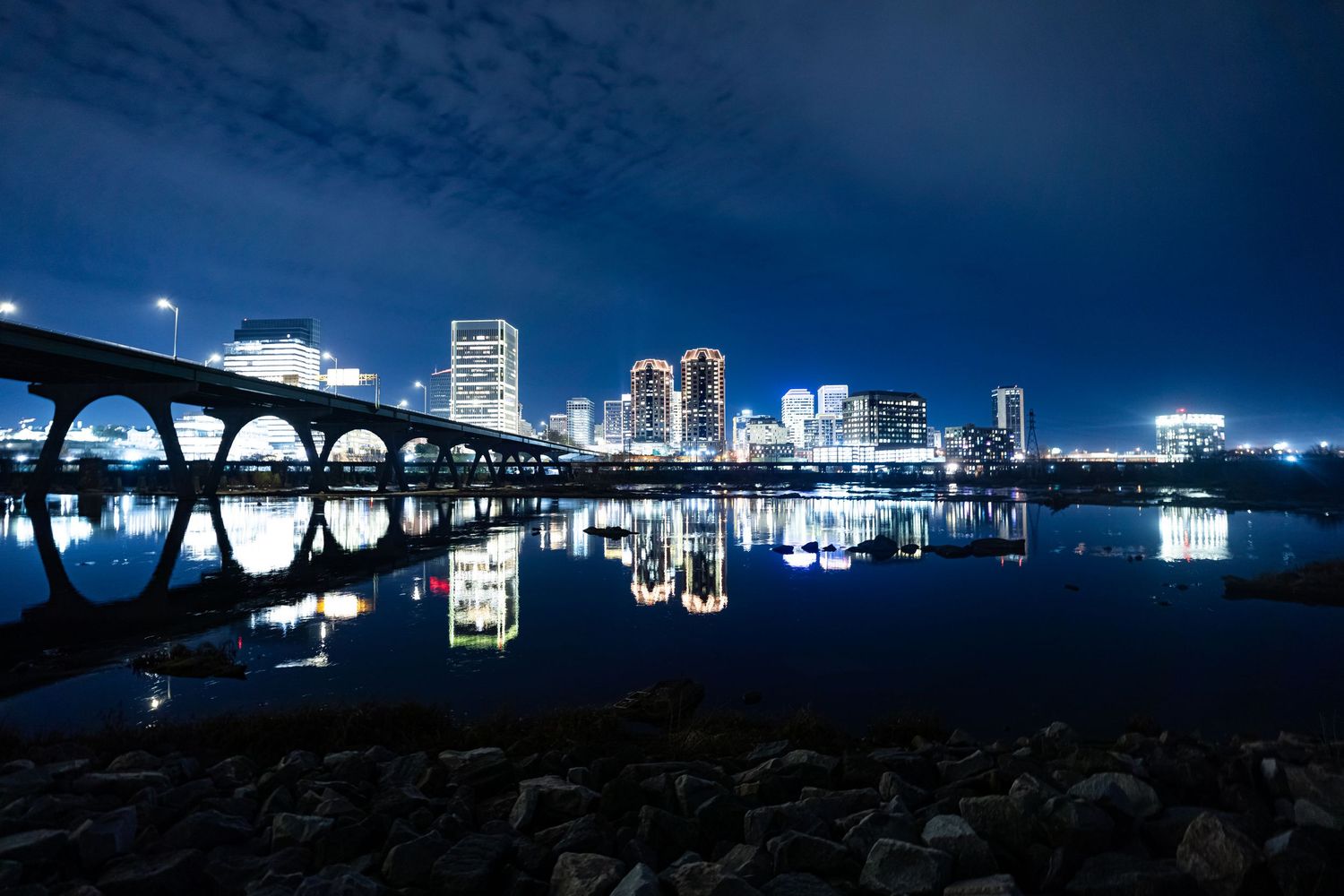Dramatic cityscape with bridge and buildings reflected in water under moody sky.