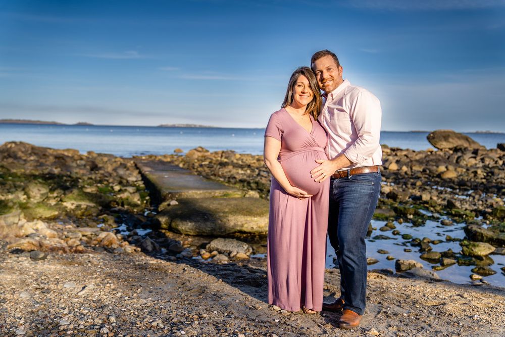 Family poses for photos along a rocky ocean shoreline at sunset with blue water and mountains in background.
