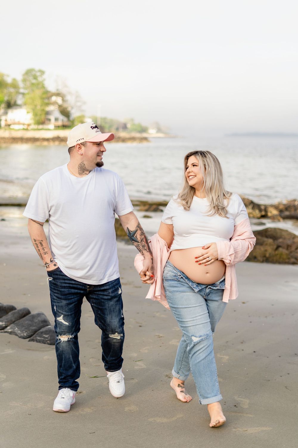 Expecting couple walks barefoot along a rocky beach during sunset while showing off her baby bump.