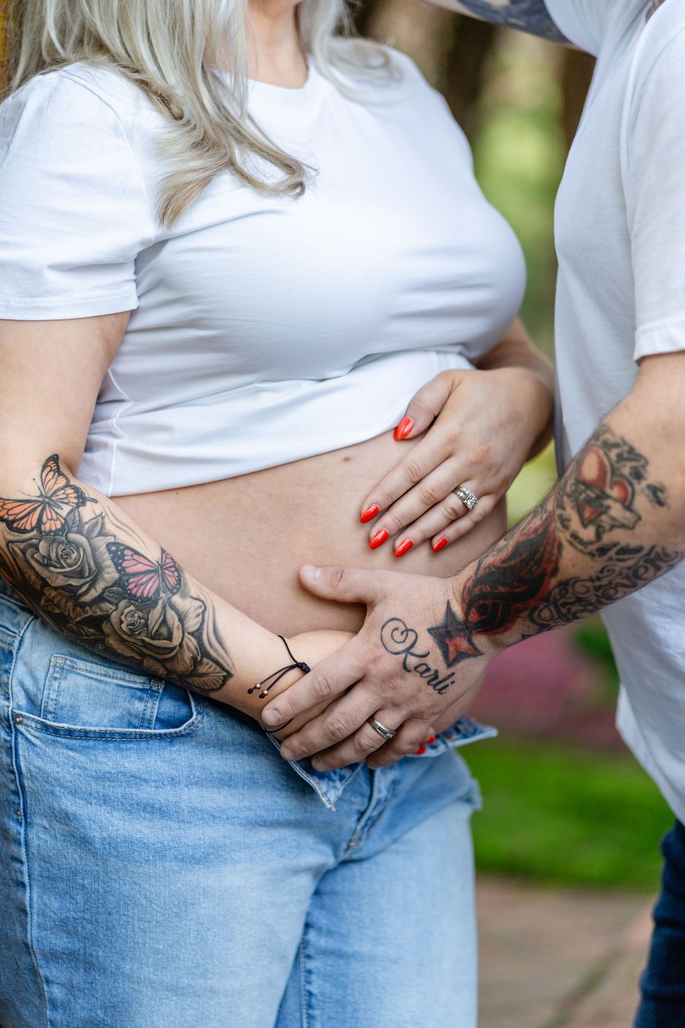 Close-up photo showing a tattooed belly in a white shirt and jeans with someone's hands resting on the stomach.
