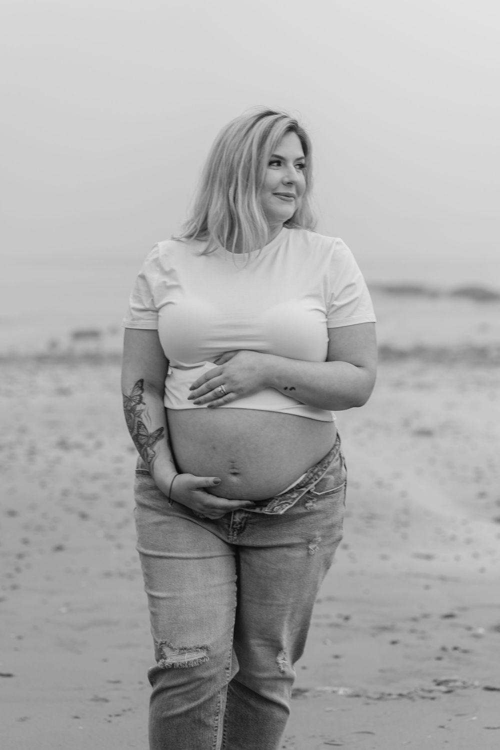 Black and white maternity photo of an expectant mother in casual clothes posing on a beach.