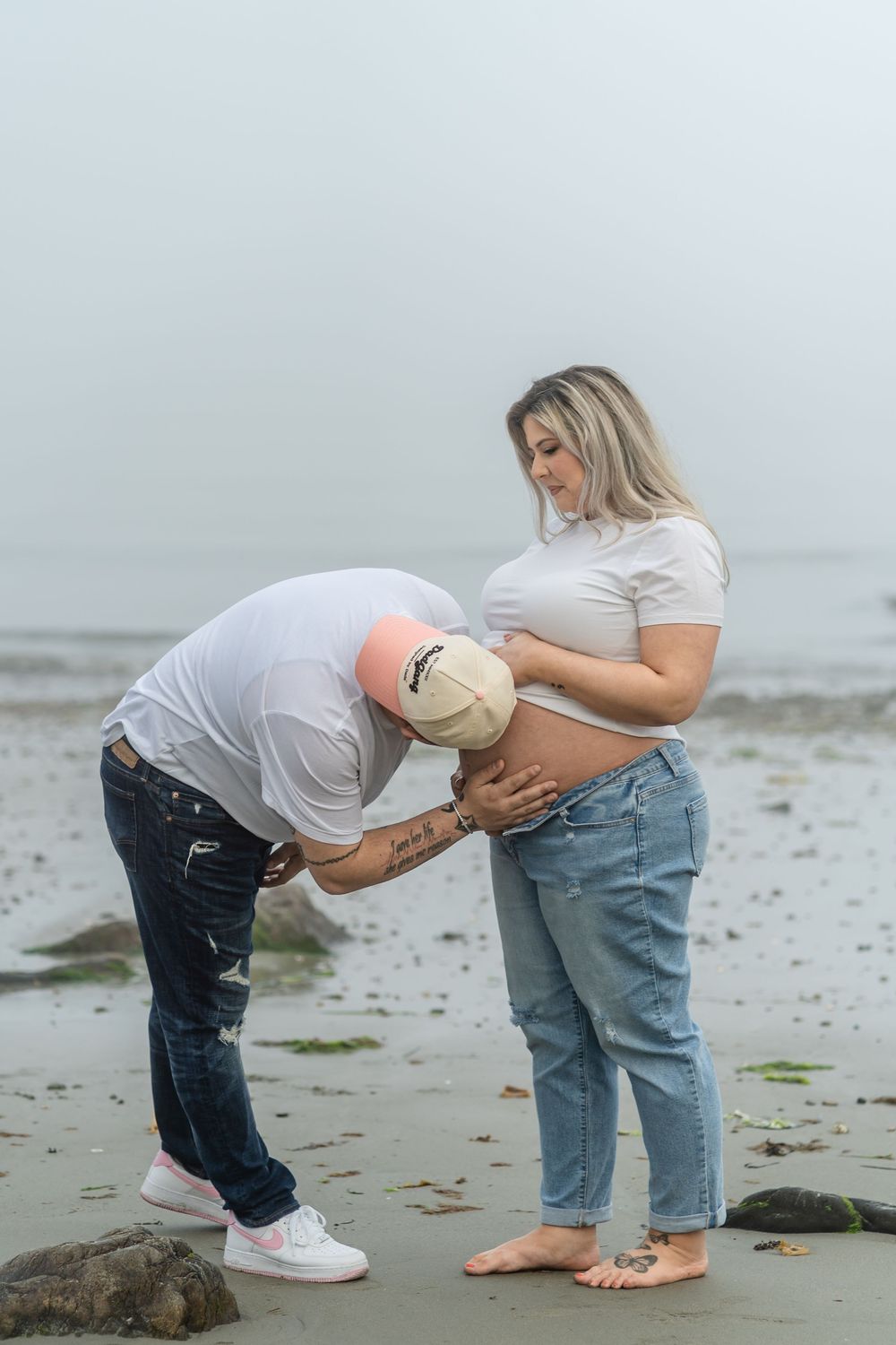 People bent over on a beach taking photos of someone's tattooed stomach against an overcast sky.