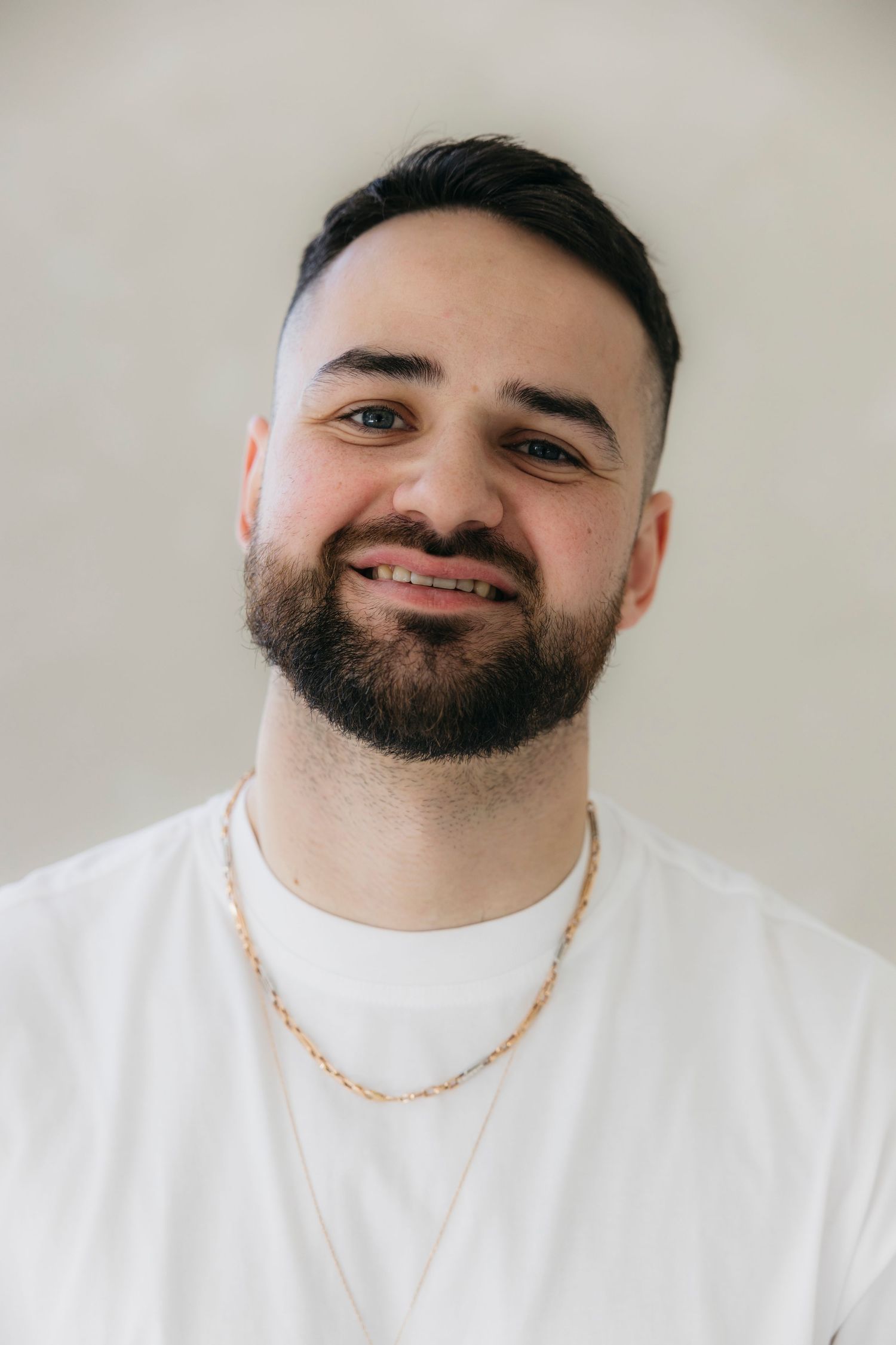 A portrait of a person in a white t-shirt and gold chain smiling against a light background.
