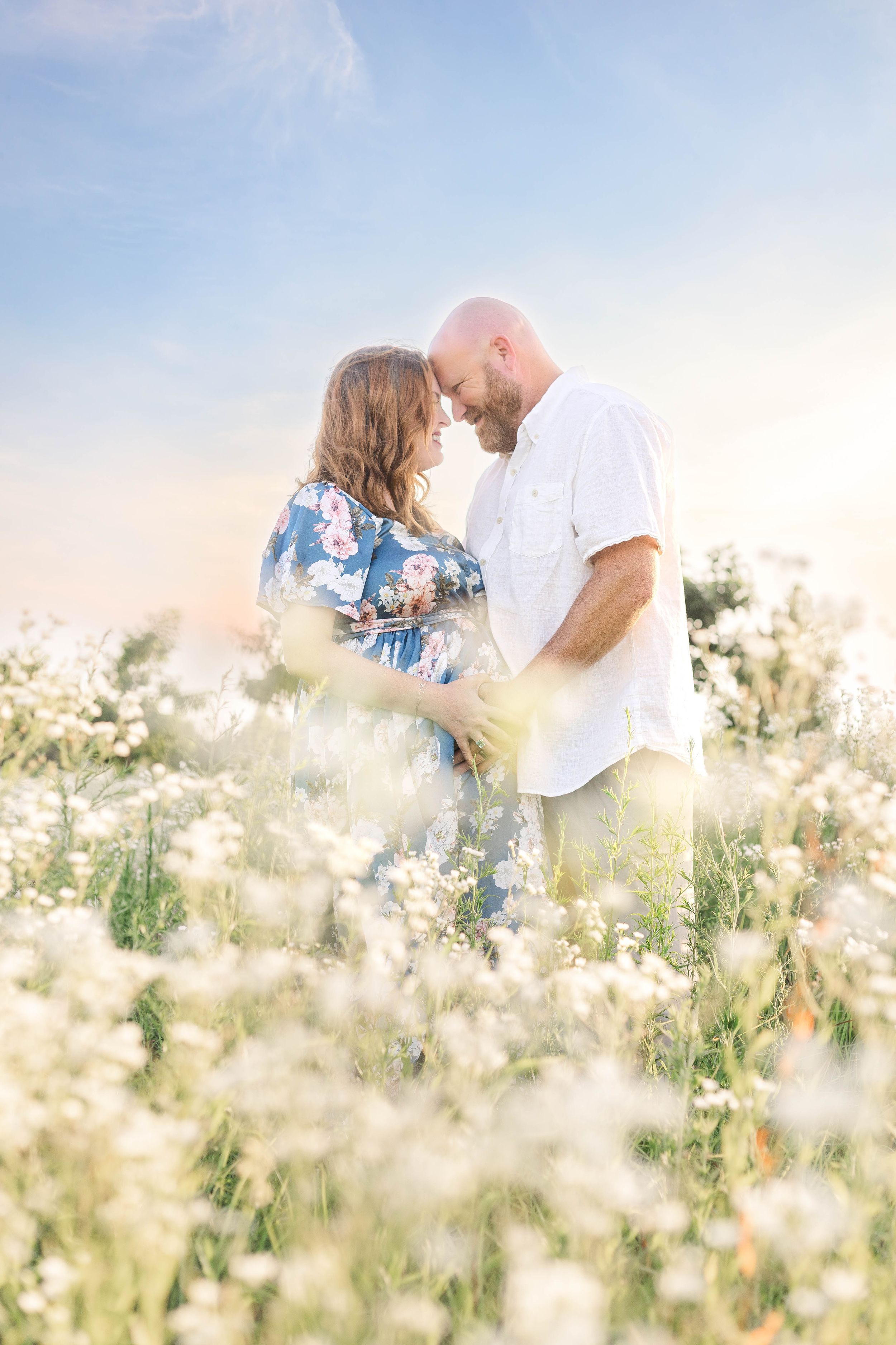 Expectant couple embracing in a field of white wildflowers, with the mother wearing a flowing blue floral maternity dress.