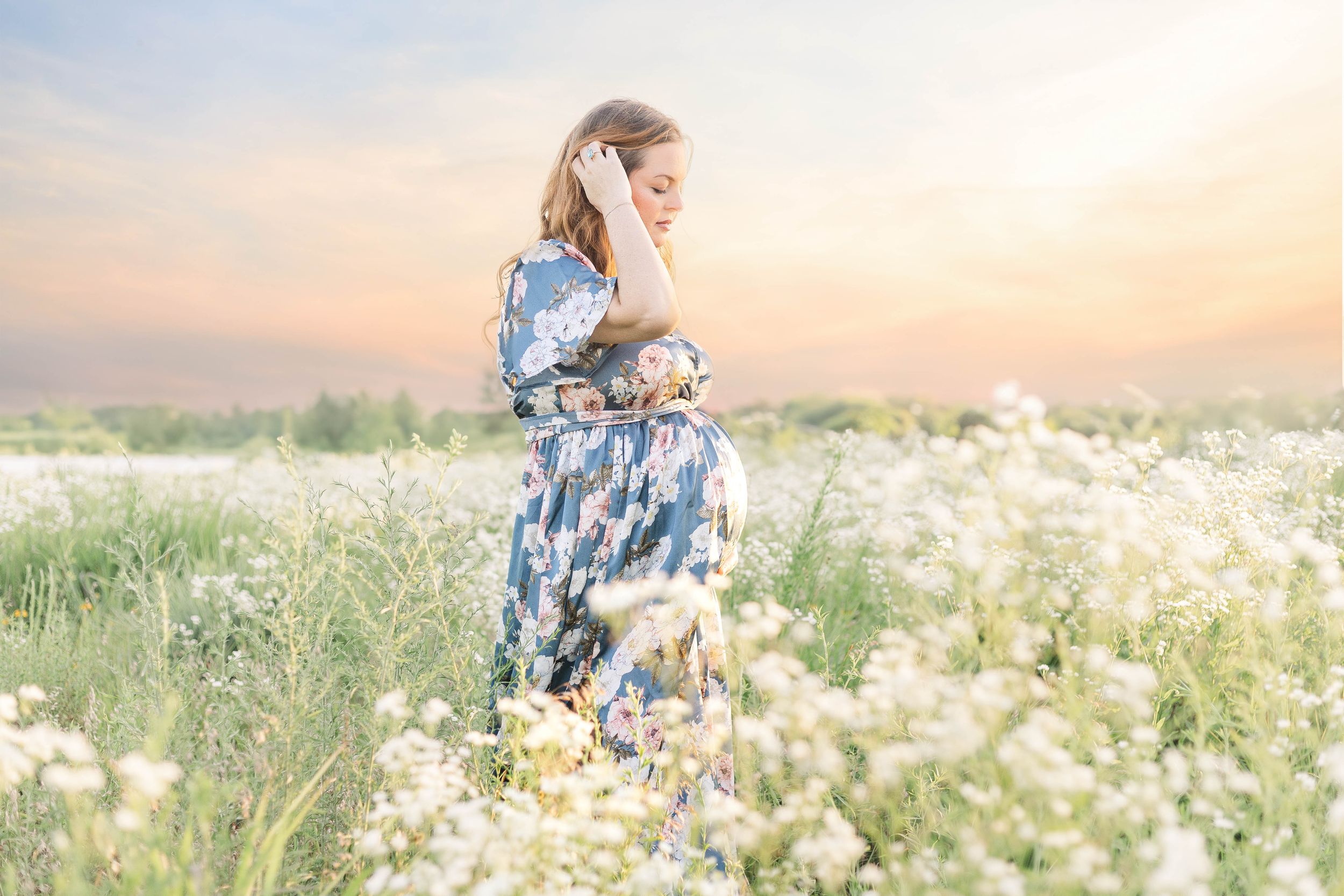 Expectant mother wearing a blue floral dress standing in a field of white wildflowers during a sunset maternity portrait session.