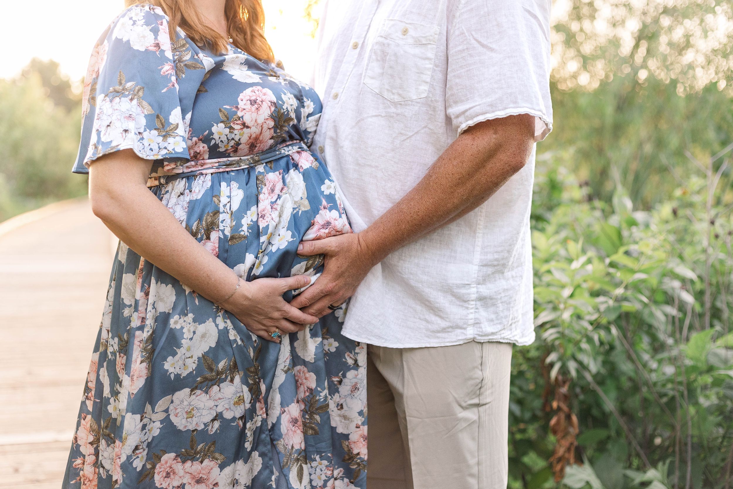 Close-up of expectant parents gently holding the mother's baby bump, with her wearing a blue floral maternity dress outdoors.