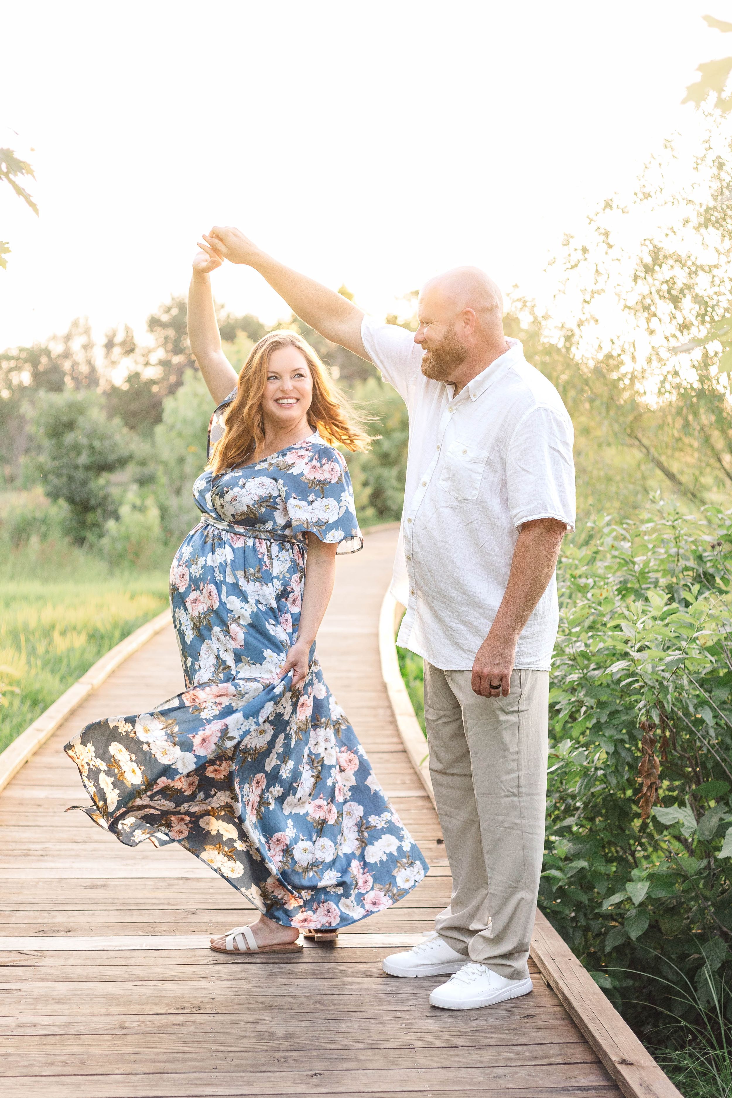 Expectant couple standing together on a wooden walkway in warm evening sunlight during a natural outdoor maternity portrait session.