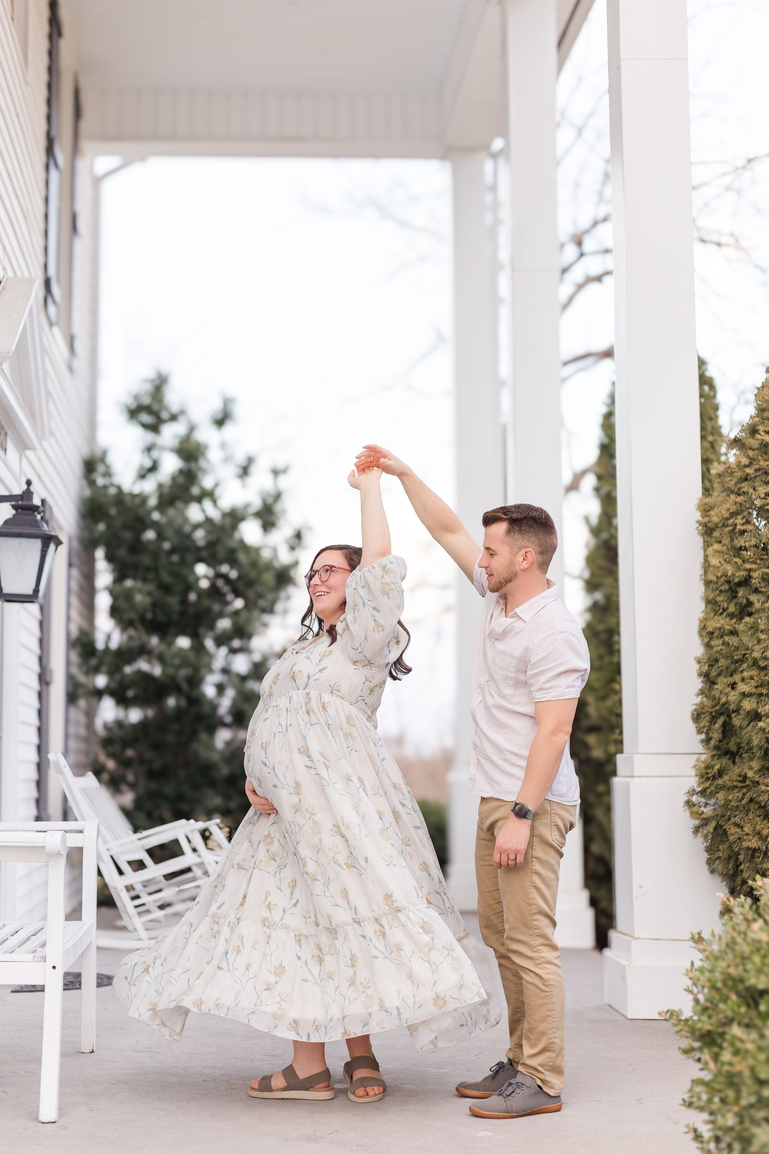 Expectant couple sharing a playful twirl beneath a covered outdoor walkway, with the mother wearing a flowing floral maternity dress.