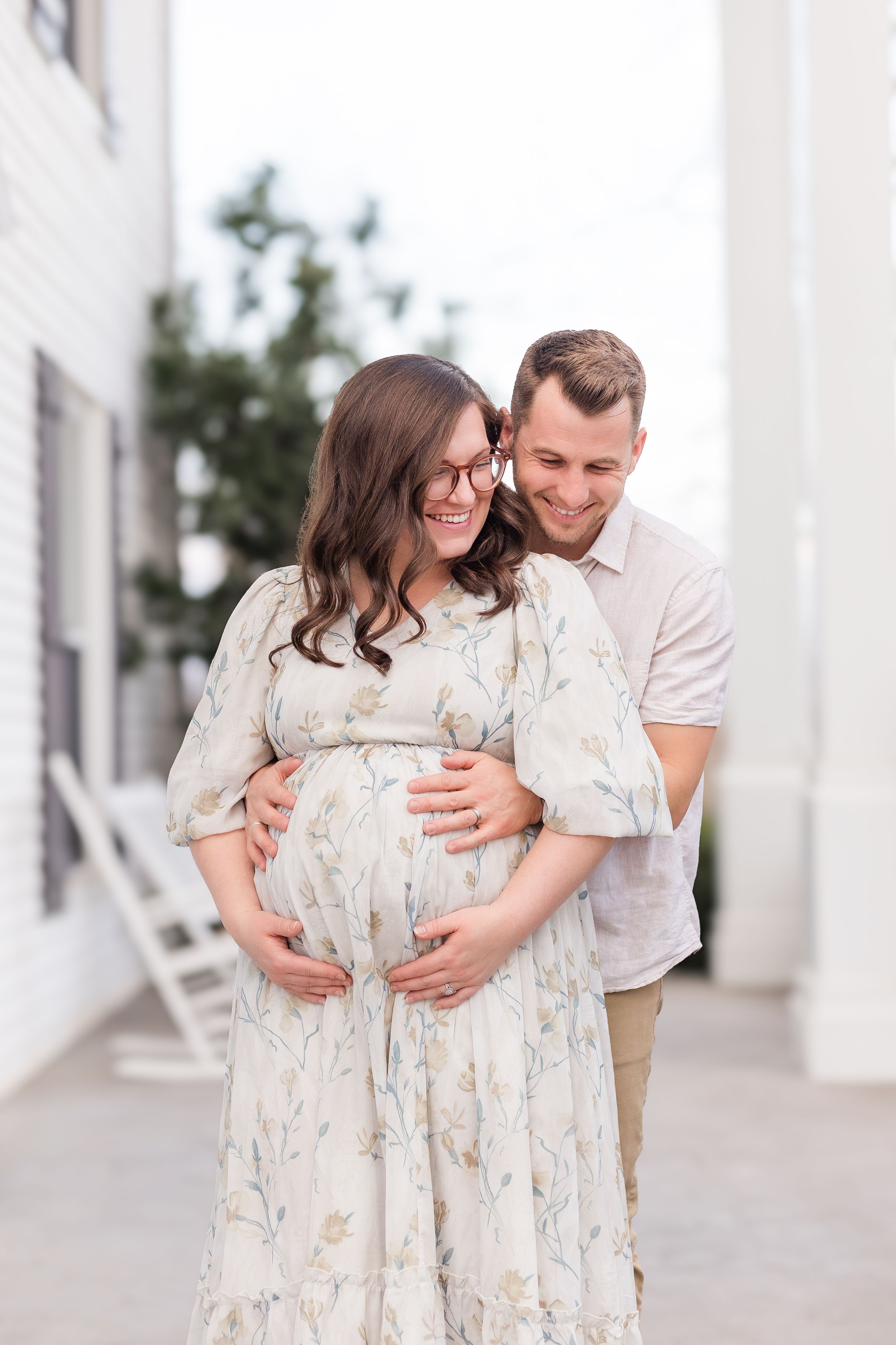 Expectant mother in a floral dress smiling as her partner embraces her from behind during an outdoor maternity photography session.