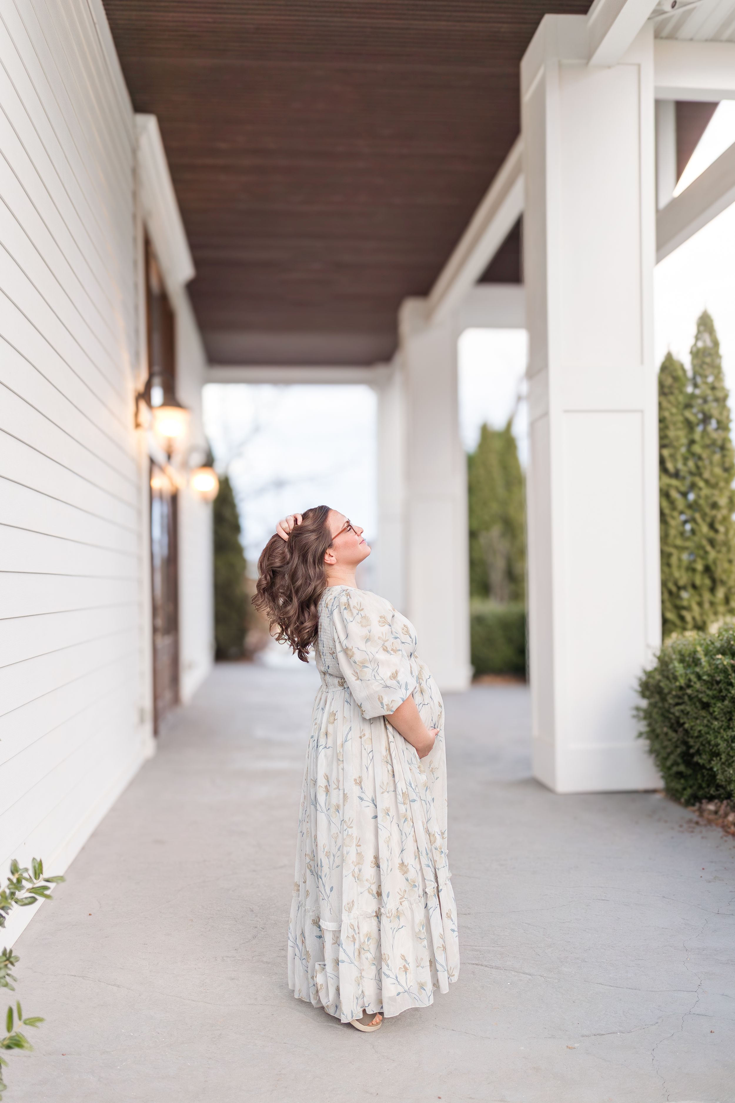Expectant mother wearing a flowing floral dress standing beneath a covered outdoor walkway during a natural maternity portrait session.