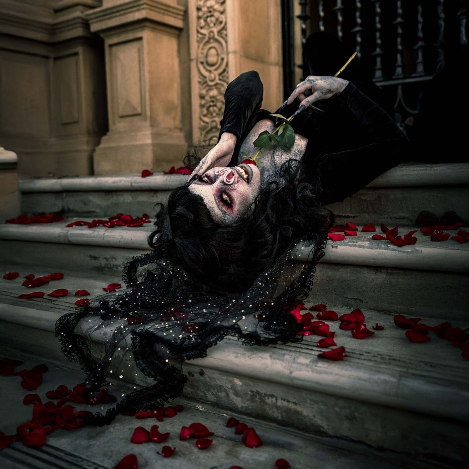 Female model in a haunting yet beautiful bat-inspired horror shoot, lying upside down on steps surrounded by rose petals.