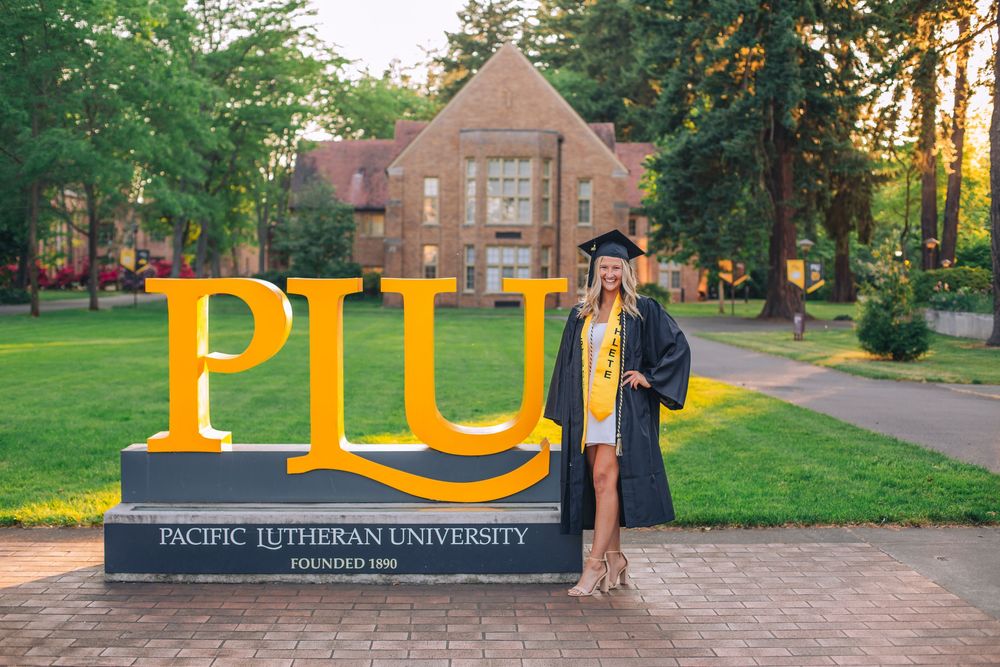 Graduate in cap and gown poses by PLU university sign during sunset. Spokane Liberty Lake Photographer Senior Photographer