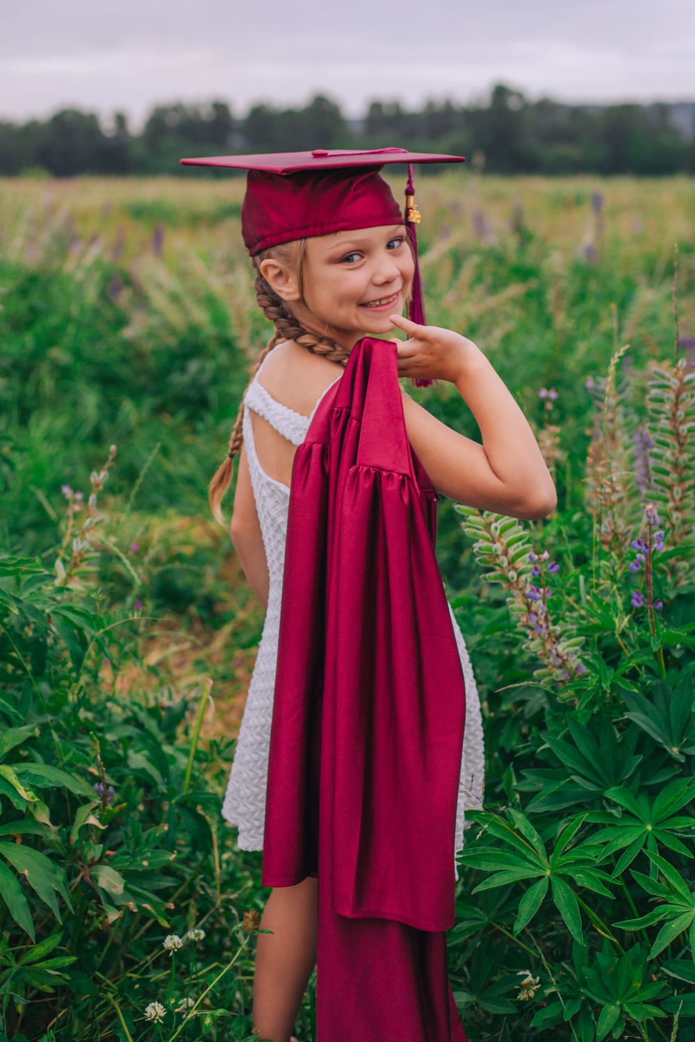 Young child in white dress poses with red graduation cap and gown in wildflower field. Spokane Liberty Lake Photographer Senior Photographer
