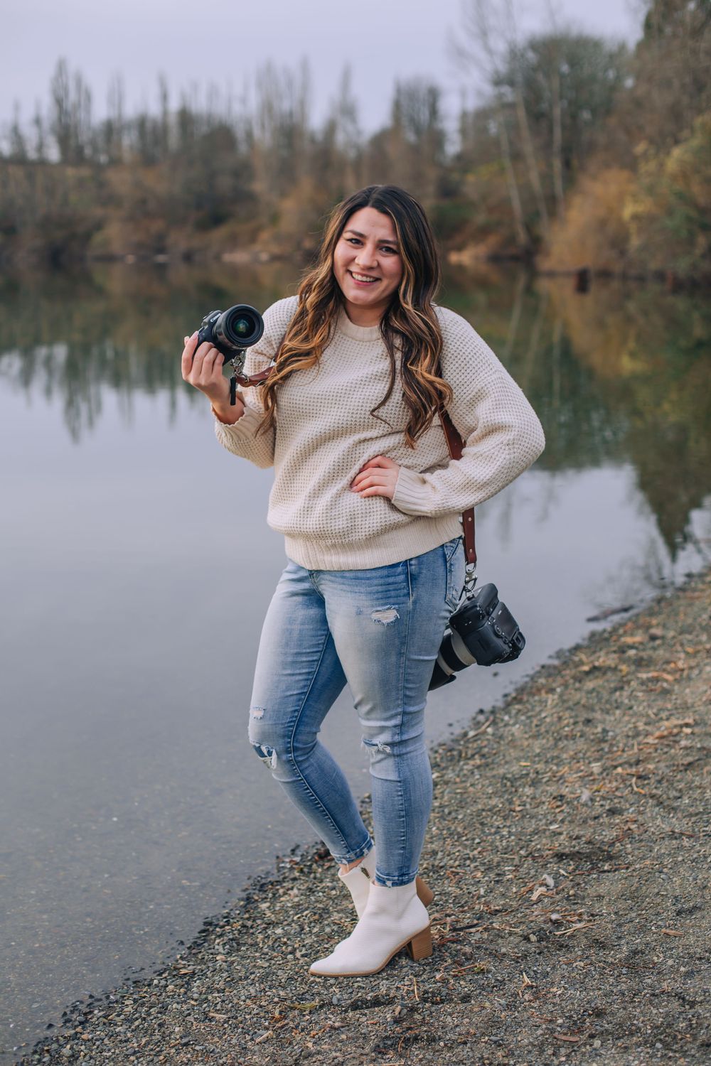 Photographer in cream sweater and jeans poses by lakeside on rocky shore. Spokane Liberty Lake Photographer Portrait Photographer