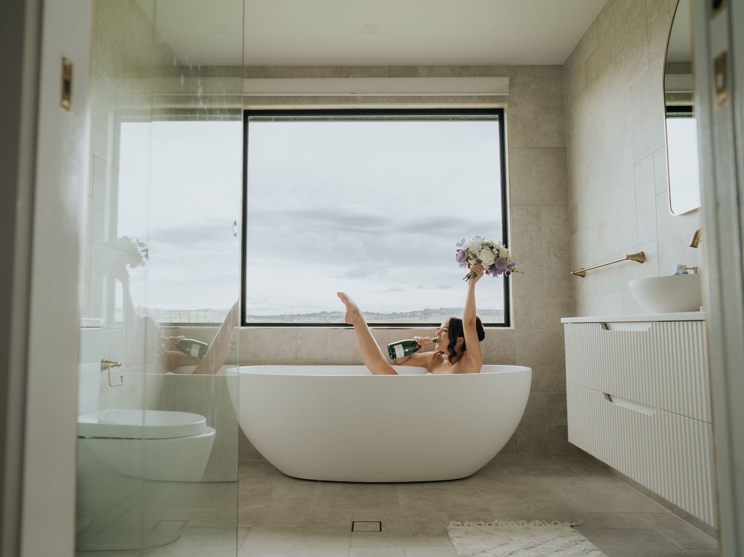 Bride relaxing in freestanding bathtub holding floral bouquet with panoramic ocean views through window.