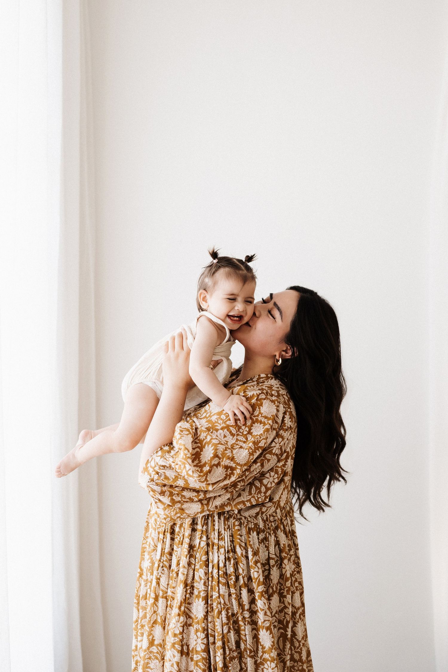 Mother in floral dress lifting and kissing baby against white wall.