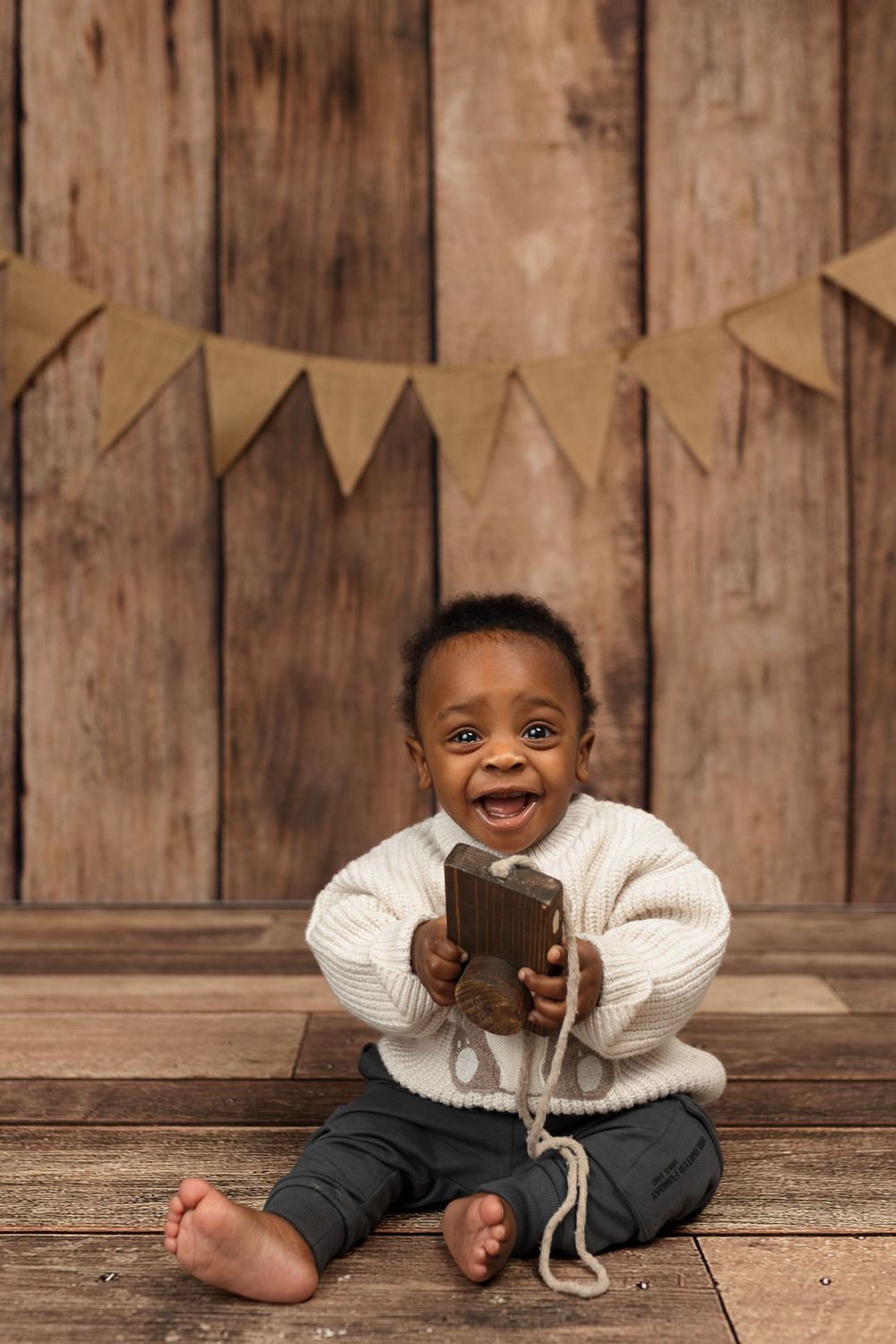 Smiling baby boy in white sweater sitting on wooden floor holding a toy phone with burlap bunting backdrop.