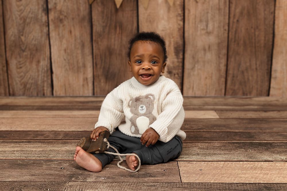 Smiling baby boy in white teddy bear sweater and jeans sits cross-legged on wood floor against rustic wooden wall.