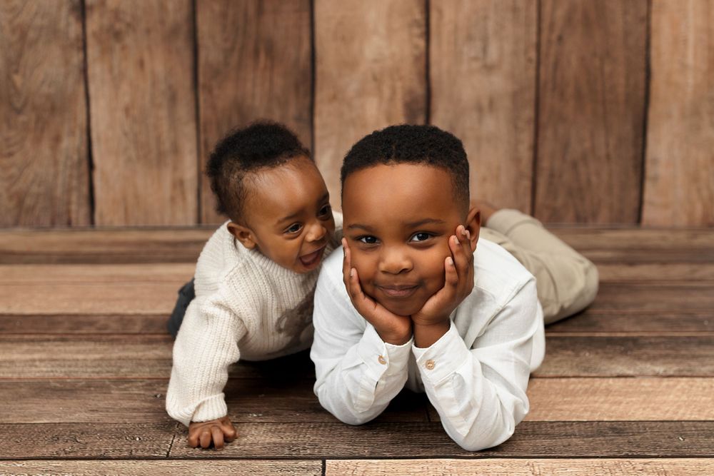 Two young brothers pose together on wooden floor, older boy lying down smiling while baby sibling crawls beside him.
