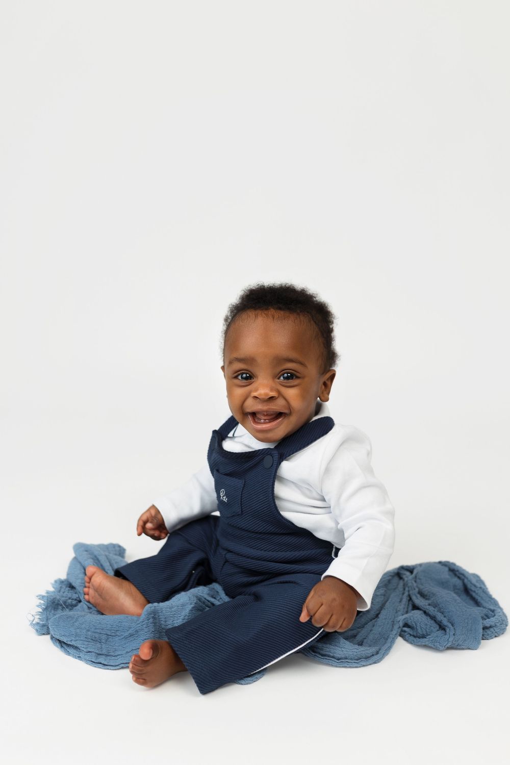 Smiling baby in navy overalls and white shirt sits on white background with blue blanket.