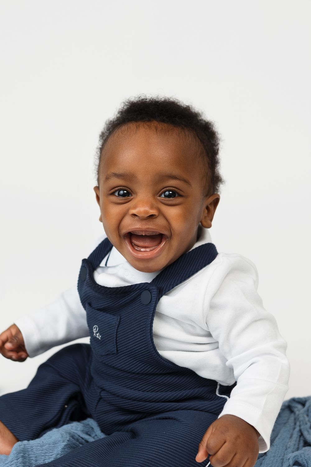 A happy baby boy in a navy blue overalls and white long-sleeve shirt smiling joyfully against a white background.