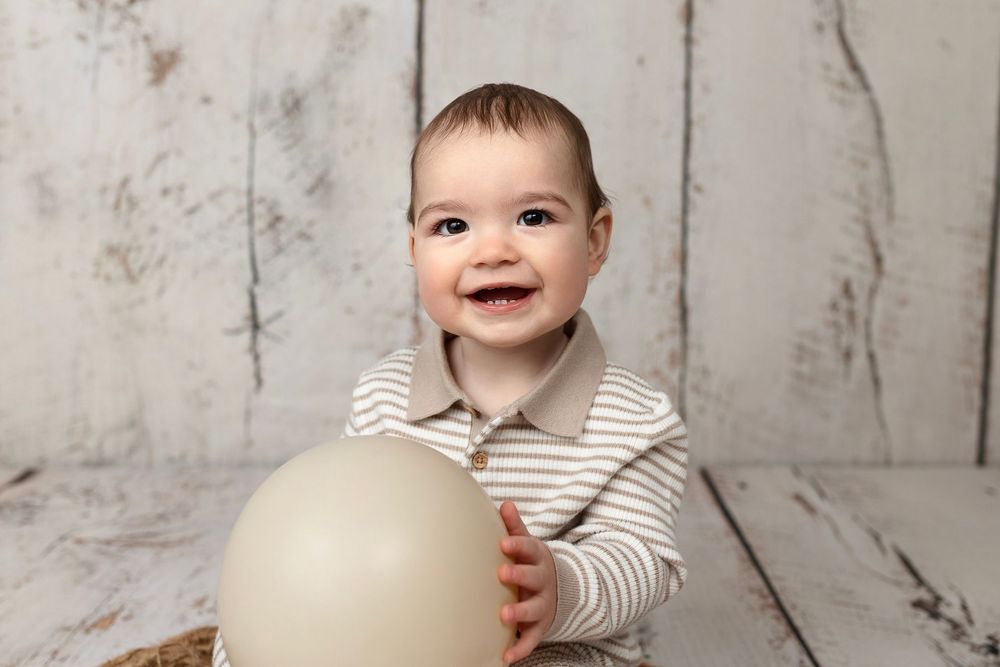 Smiling baby boy in striped outfit holding a beige balloon against a rustic white wood backdrop.
