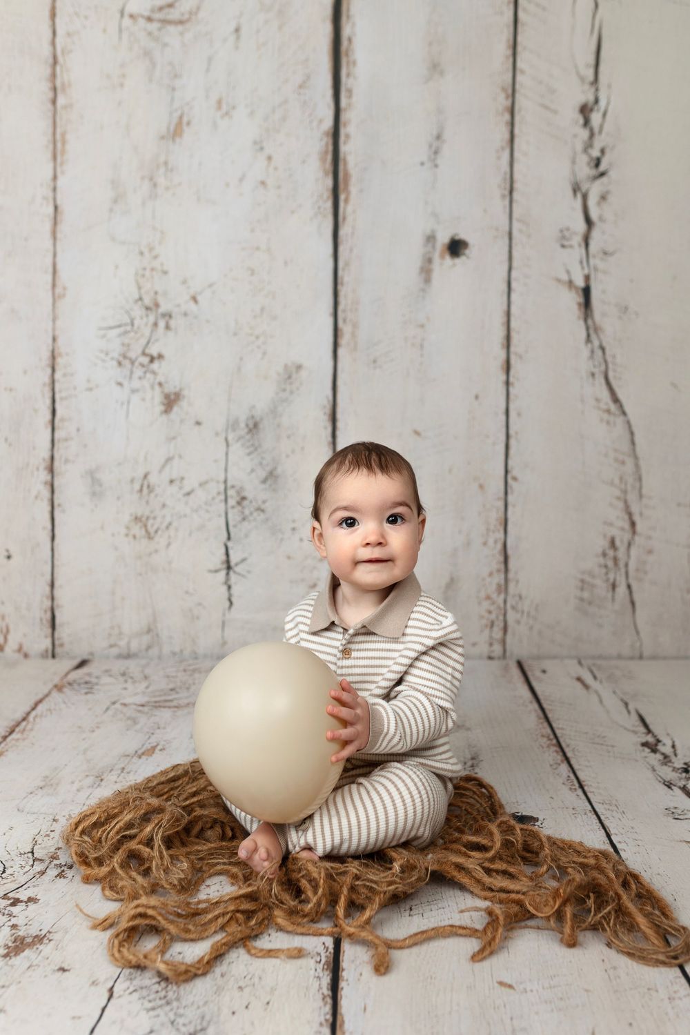 Baby in striped onesie sitting on jute mat holding a beige balloon against a rustic white wood backdrop.