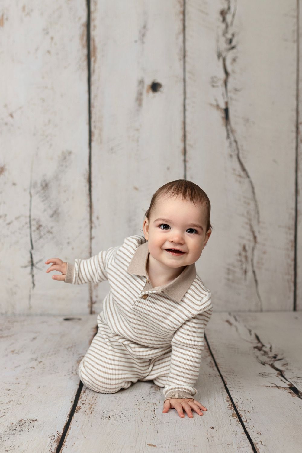 Smiling baby in striped romper sitting on white rustic wood floor against distressed white wood backdrop.