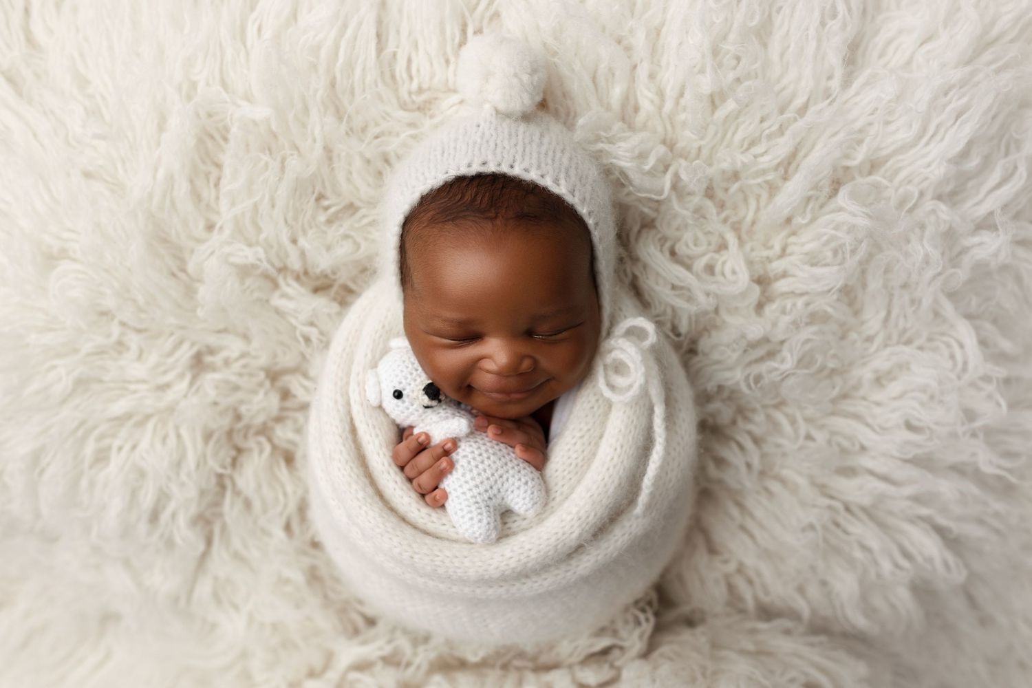 Peaceful newborn wearing knit hat sleeping in cream wrap on fluffy gray backdrop.